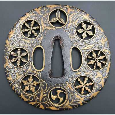 A metal tsuba (sword guard) features engraved leaves (foliage), flowers (blossoms), and circular family crests (mon). The surface exhibits gold coloring on the engravings, contrasting with the darker metal background."