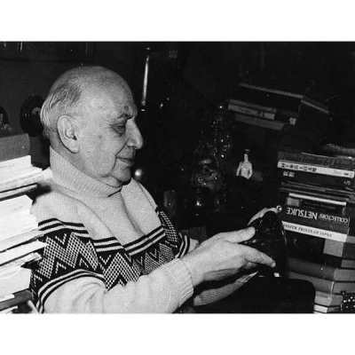 An old man examining a dark-colored, patterned object (possibly camera). He is seated, surrounded by piles of books. His sweater features a geometric pattern."