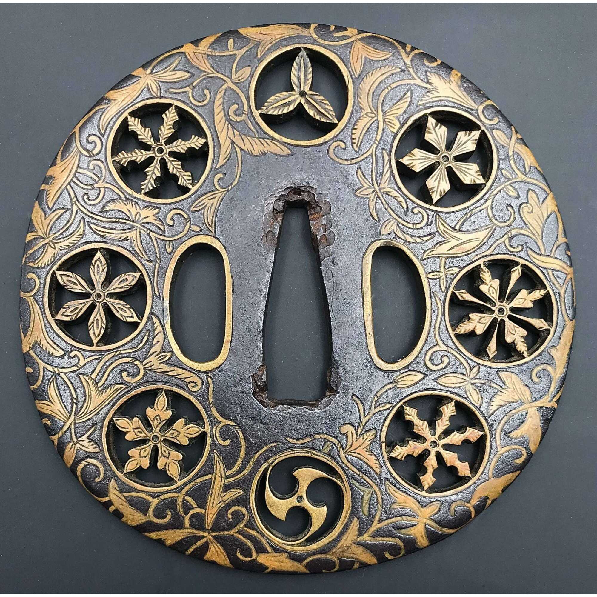 A metal tsuba (sword guard) features engraved leaves (foliage), flowers (blossoms), and circular family crests (mon). The surface exhibits gold coloring on the engravings, contrasting with the darker metal background."